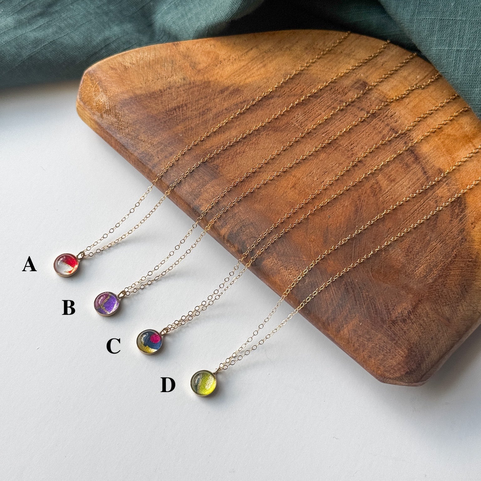 Four delicate gold-tone necklaces with small round gemstone pendants labeled A, B, C, and D, arranged on a curved wooden tray with a dark teal fabric backdrop.