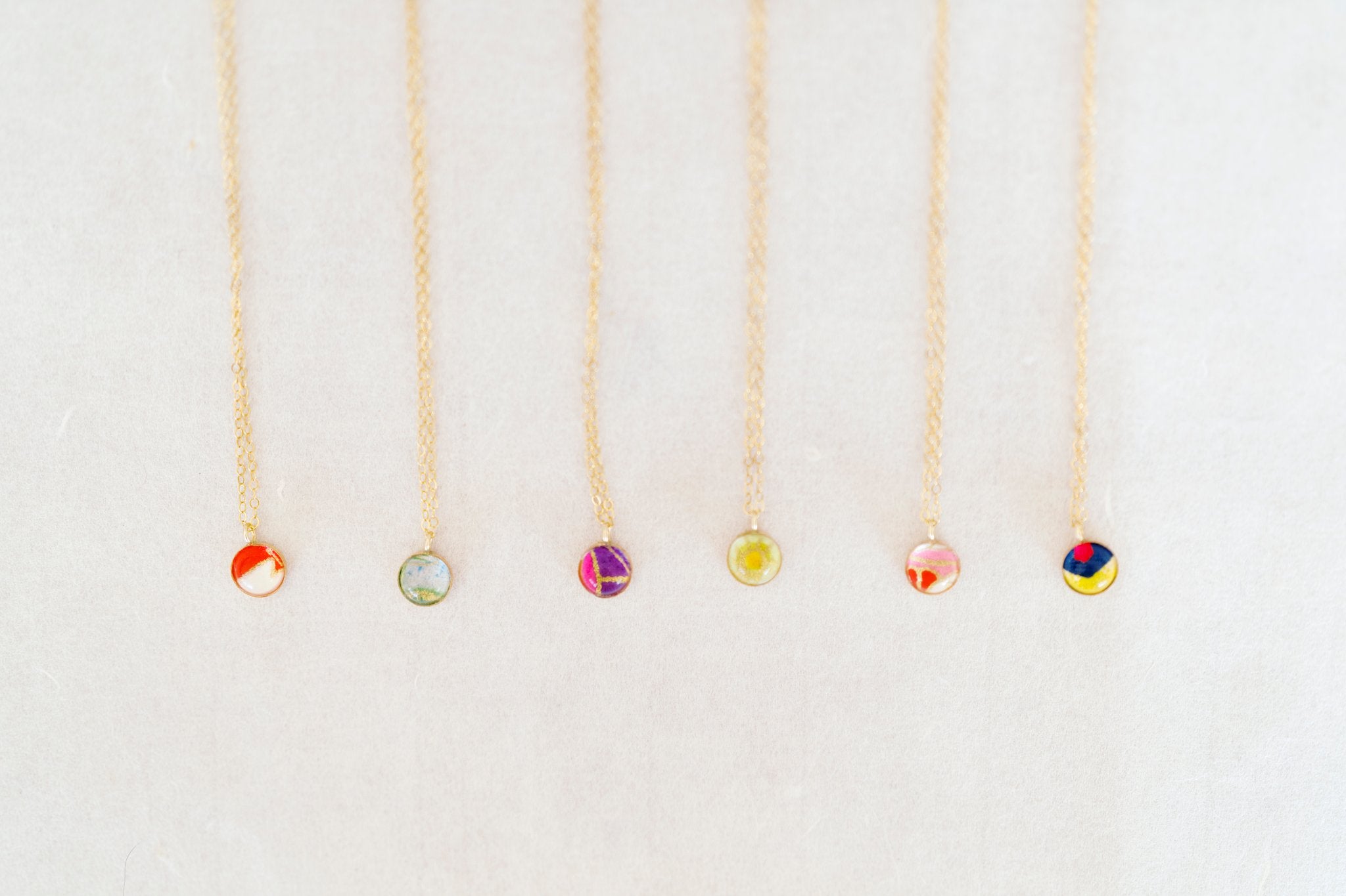 Six delicate gold necklaces with small round glass bead pendants in various colors, arranged vertically on a light beige textured background.