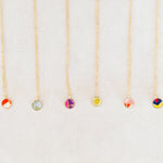 Six delicate gold necklaces with small round glass bead pendants in various colors, arranged vertically on a light beige textured background.