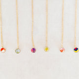 Six delicate gold necklaces with small round glass bead pendants in various colors, arranged vertically on a light beige textured background.