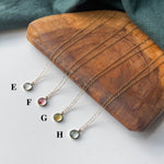 Four delicate gold-tone necklaces with small round gemstone pendants labeled E, F, G, and H, arranged on a curved wooden tray with a dark teal fabric backdrop.