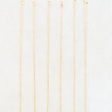 Six delicate gold chain necklaces with small, colorful circular pendants are arranged vertically in a row on a clean white background.