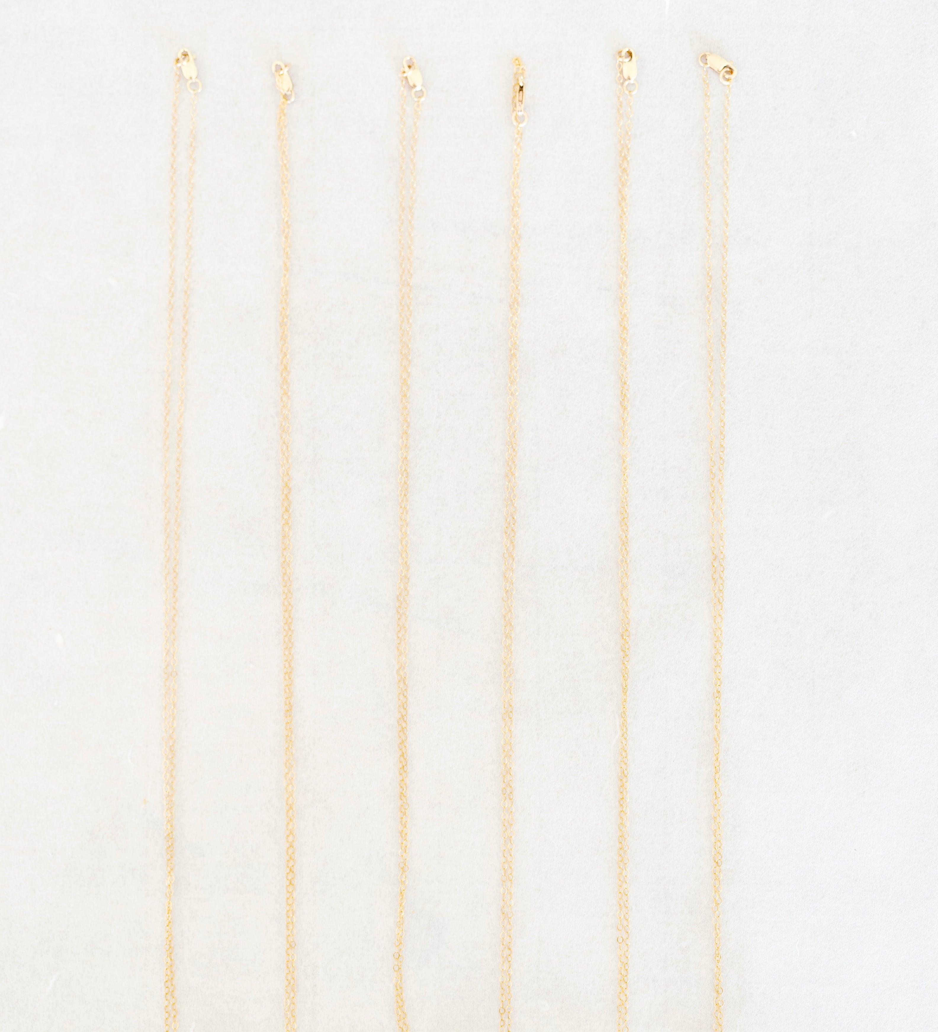 Six delicate gold chain necklaces with small, colorful circular pendants are arranged vertically in a row on a clean white background.