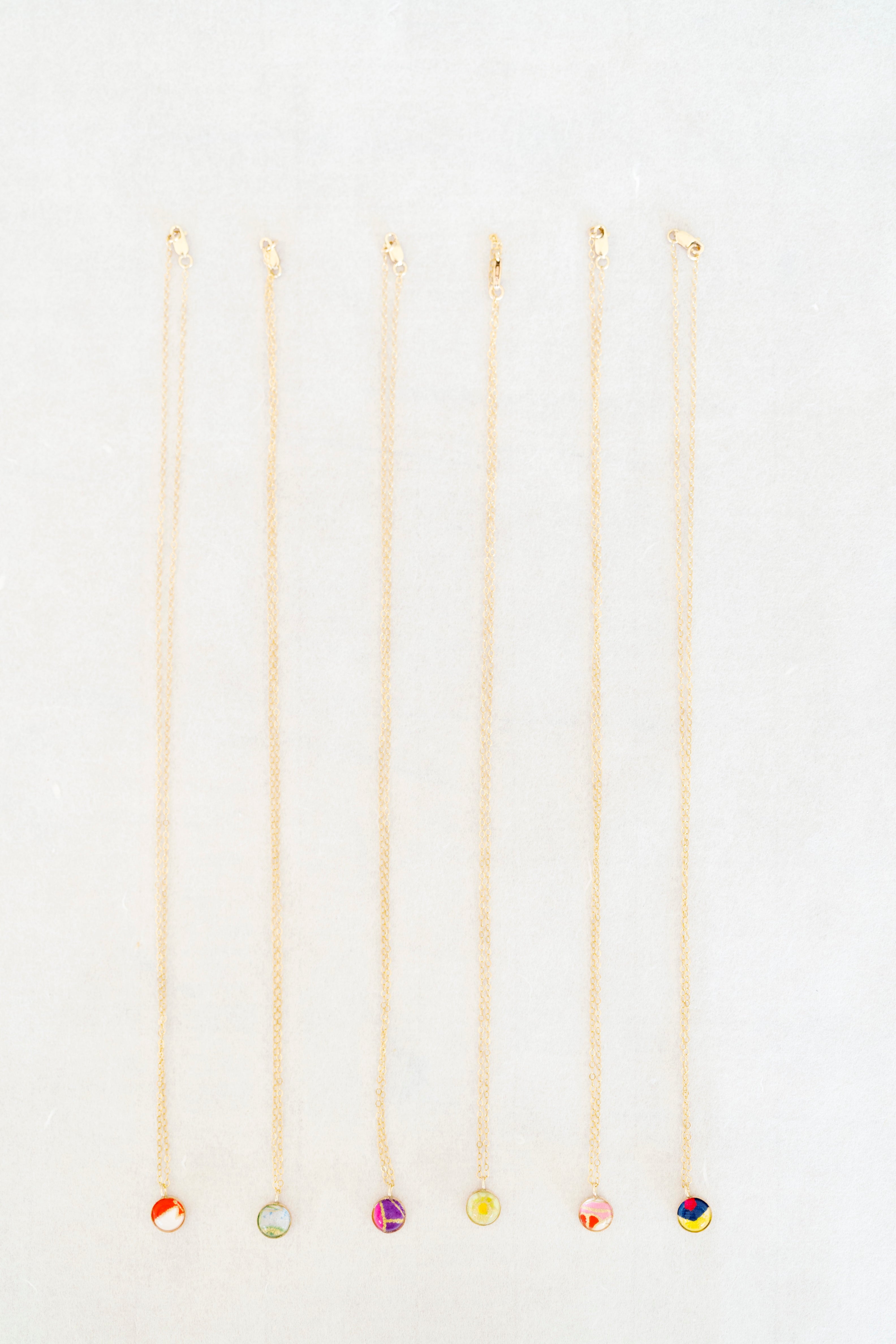 Six delicate gold chain necklaces with small, colorful circular pendants are arranged vertically in a row on a clean white background.