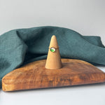 A delicate gold ring with a small round green and white cabochon displayed on a wooden cone stand, set on a rustic wooden board with a draped teal fabric background.