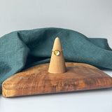 A delicate gold ring with a small clear stone is displayed on a wooden cone stand, set on a rustic wooden board with a draped teal linen cloth against a light gray background.