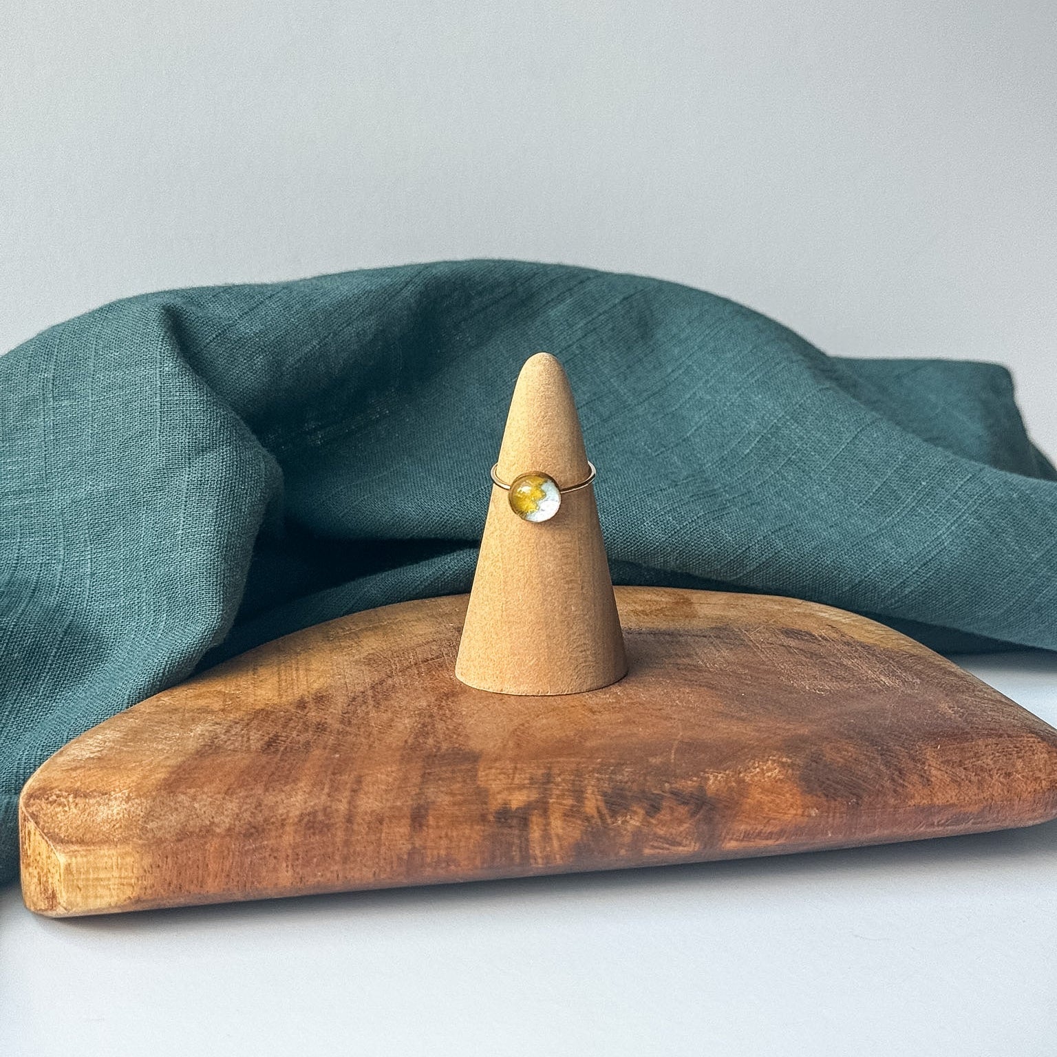 A delicate gold ring with a small clear stone is displayed on a wooden cone stand, set on a rustic wooden board with a draped teal linen cloth against a light gray background.