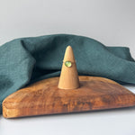 A delicate gold ring with a small clear stone is displayed on a wooden cone stand, set on a rustic wooden board with a draped teal linen cloth against a light gray background.