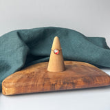 A delicate gold ring with a small clear stone is displayed on a wooden cone stand, set on a rustic wooden board with a draped teal linen cloth against a light gray background.