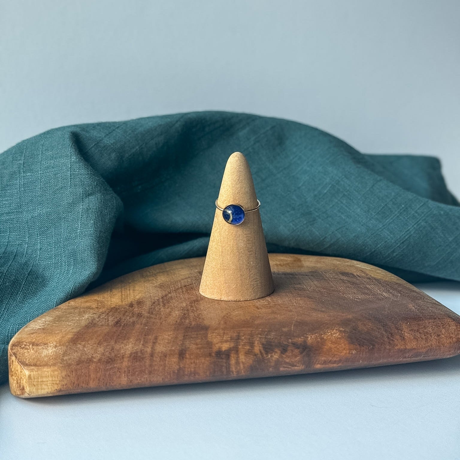 A delicate gold ring with a small clear stone is displayed on a wooden cone stand, set on a rustic wooden board with a draped teal linen cloth against a light gray background.