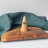A delicate gold ring with a small black stone is displayed on a wooden cone stand, set on a rustic wooden board with a draped teal linen cloth against a light gray background.
