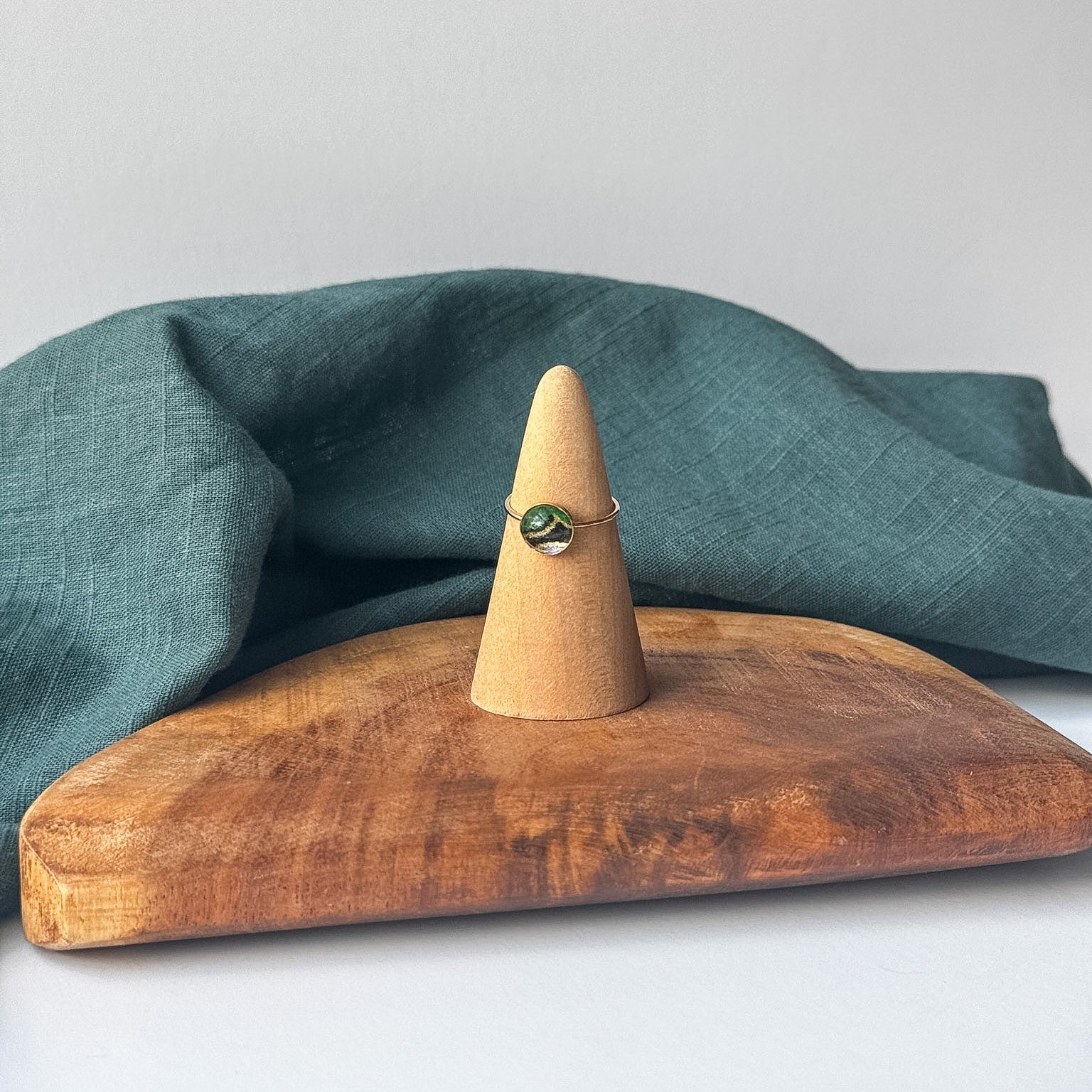 A delicate gold ring with a small black stone is displayed on a wooden cone stand, set on a rustic wooden board with a draped teal linen cloth against a light gray background.