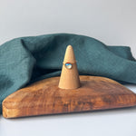 A delicate gold ring with a small clear stone is displayed on a wooden cone stand, set on a rustic wooden board with a draped teal linen cloth against a light gray background.