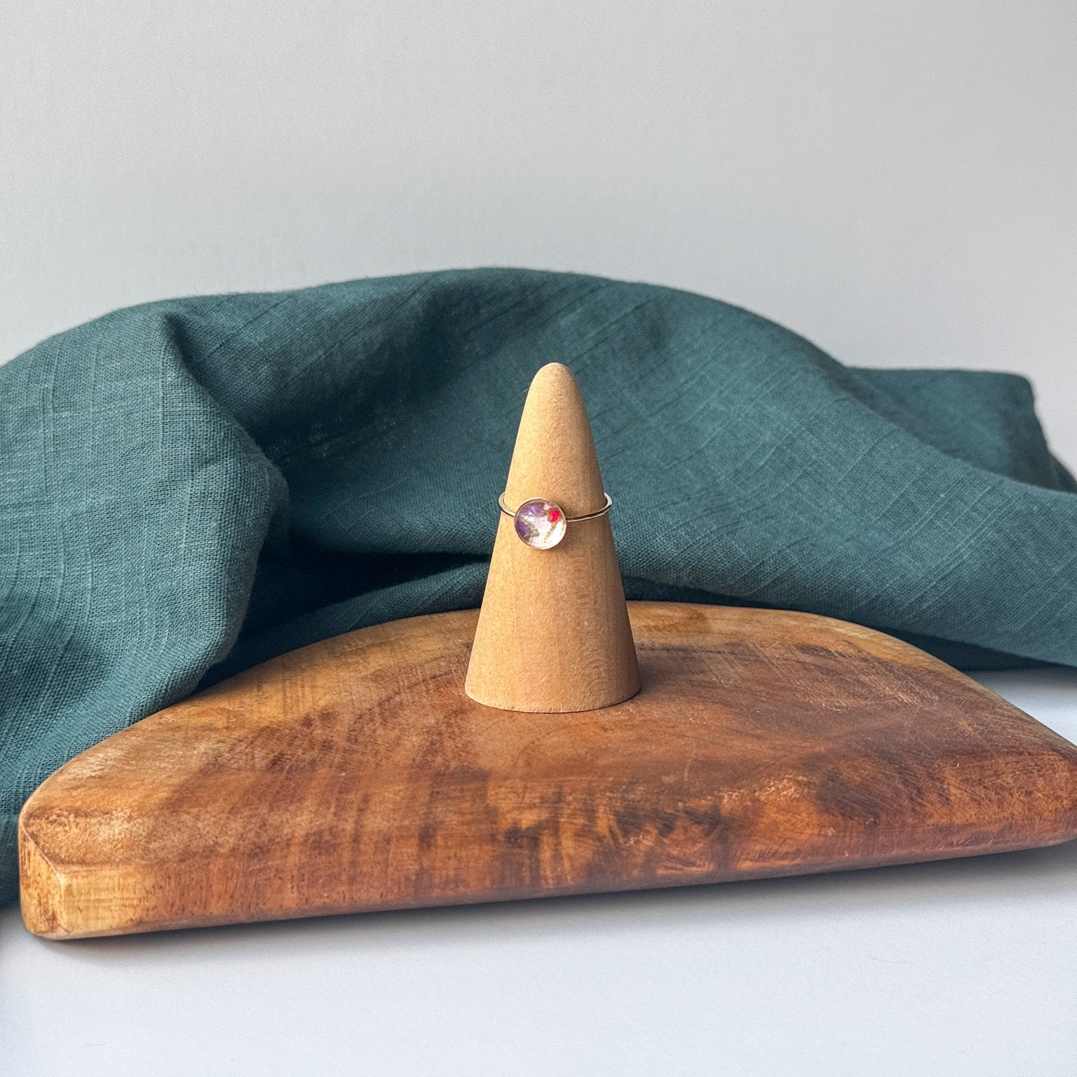 A delicate gold ring with a small pink stone is displayed on a wooden cone stand, set on a rustic wooden board with a draped teal linen cloth against a light gray background.