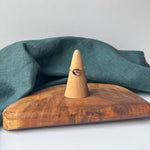 A delicate gold ring with a small purple stone is displayed on a wooden cone stand, set on a rustic wooden board with a draped teal linen cloth against a light gray background.
