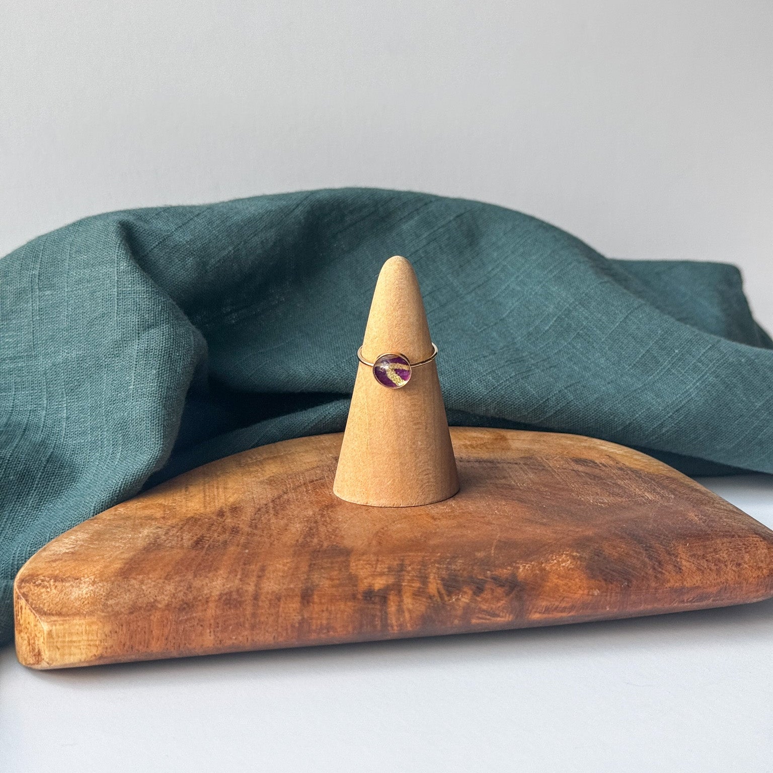 A delicate gold ring with a small purple stone is displayed on a wooden cone stand, set on a rustic wooden board with a draped teal linen cloth against a light gray background.