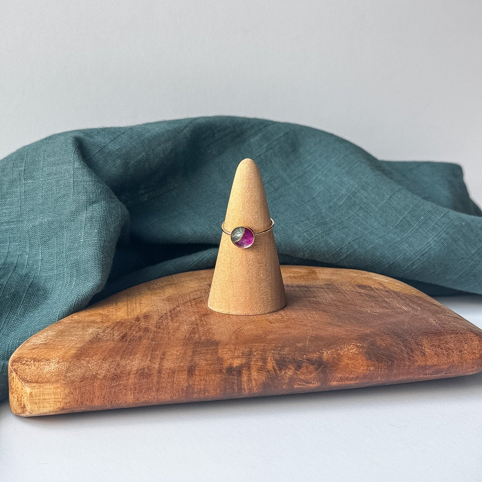 A delicate gold ring with a small purple stone is displayed on a wooden cone stand, set on a rustic wooden board with a draped teal linen cloth against a light gray background.