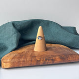 A delicate gold ring with a small blue stone is displayed on a wooden cone stand, set on a rustic wooden board with a draped teal linen cloth against a light gray background.