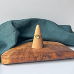 A delicate gold ring with a small green stone is displayed on a wooden cone stand, set on a rustic wooden board with a draped teal linen cloth against a light gray background.