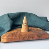 A delicate gold ring with a small green stone is displayed on a wooden cone stand, set on a rustic wooden board with a draped teal linen cloth against a light gray background.