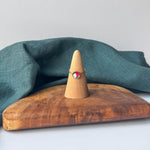 A delicate gold ring with a small red and white stone is displayed on a wooden cone stand, set on a rustic wooden board with a draped teal linen cloth against a light gray background.