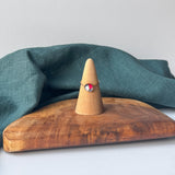 A delicate gold ring with a small red and white stone is displayed on a wooden cone stand, set on a rustic wooden board with a draped teal linen cloth against a light gray background.