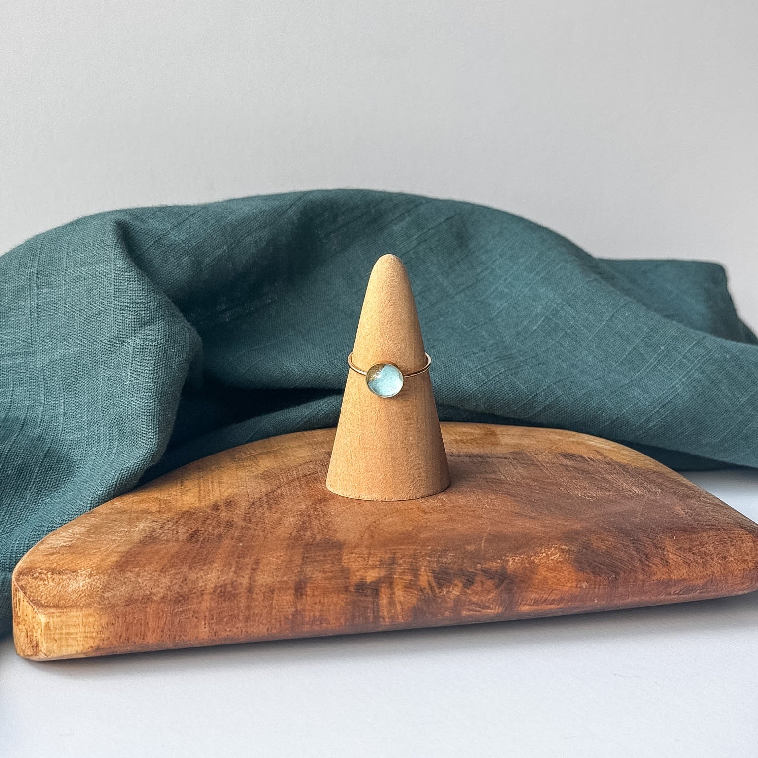 A delicate gold ring with a small blue stone is displayed on a wooden cone stand, set on a rustic wooden board with a draped teal linen cloth against a light gray background.