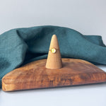 A delicate gold ring with a small yellow stone is displayed on a wooden cone stand, set on a rustic wooden board with a draped teal linen cloth against a light gray background.