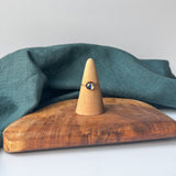 A delicate gold ring with a small navy stone is displayed on a wooden cone stand, set on a rustic wooden board with a draped teal linen cloth against a light gray background.