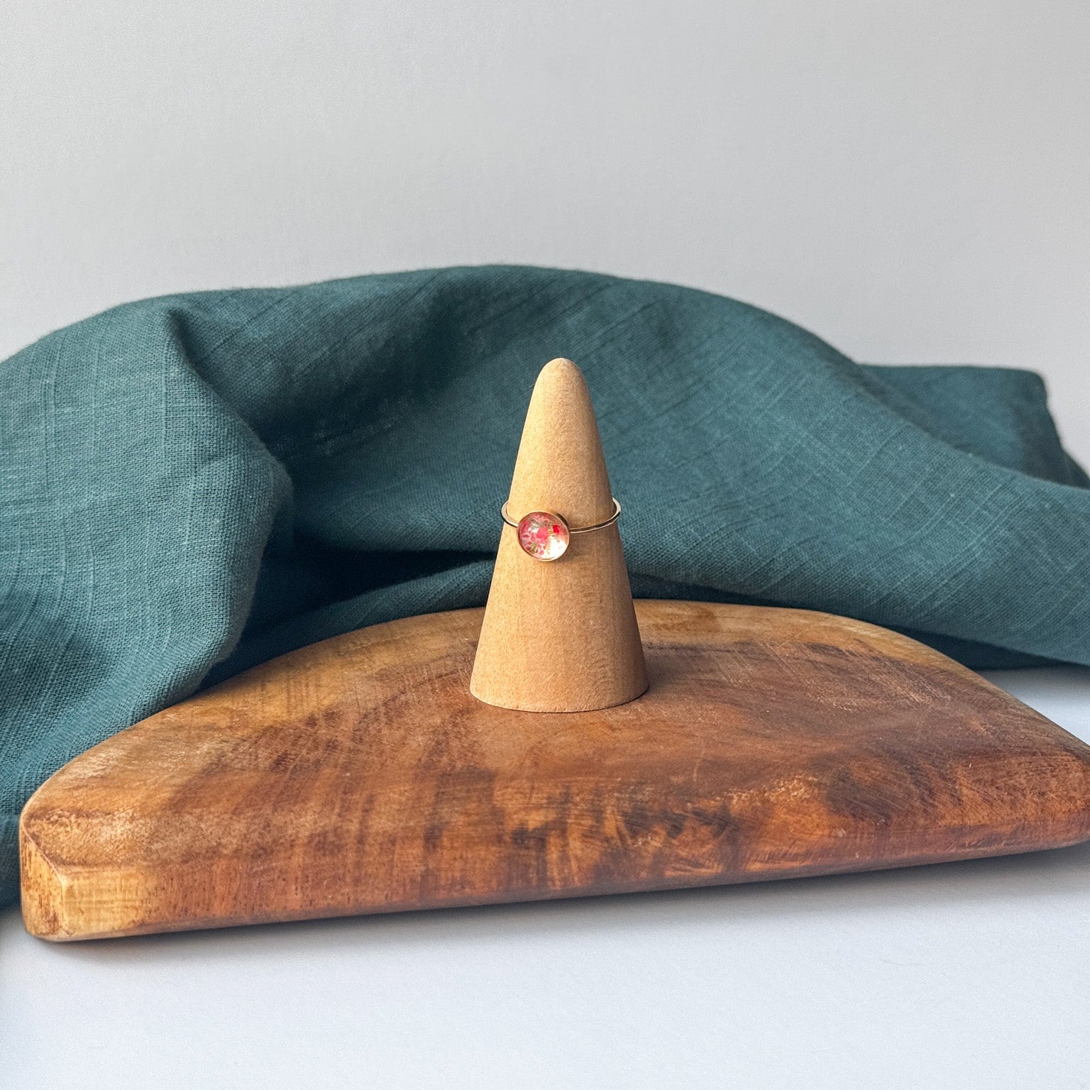 A delicate gold ring with a small pink stone is displayed on a wooden cone stand, set on a rustic wooden board with a draped teal linen cloth against a light gray background.